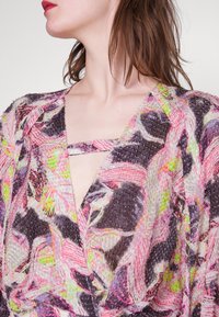 Femme à la peau claire et aux cheveux châtain moyen porte un haut texturé coloré avec des motifs abstraits rose, violet, vert et noir et une bretelle à décolleté profond.