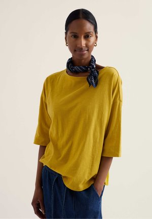 Žena s vlasmi zviazanými dozadu, nosiaca zlaté náušnice kruhy, žltú voľnú košeľu, modré nohavice a námornícky vzorovaný šál okolo krku, stojaca na svetlom pozadí.