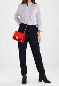 Červená prošívaná kabelka se zlatým zapínáním, nošená přes rameno. Modro-bílá pruhovaná košile, tmavé proužkované kalhoty, černé kotníkové boty.