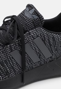 Svart atletisk sko med en texturerad knit-ovandel, tre mattsvarta ränder och en mörk gummisula. Ikonisk logotyp på tungan.