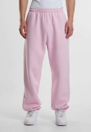 Person, die lockere hellrosa Jogginghose mit elastischem Bund und weißen Sneakers trägt, steht vor einem schlichten hellen Hintergrund.