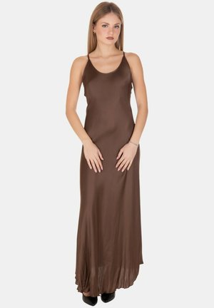 Donna con capelli lisci che indossa un abito maxi senza maniche in satin marrone e scarpe nere a punta, in piedi con le mani tese davanti.