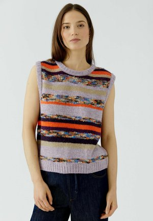 Frau mit langem braunem Haar, die einen bunten gestreiften gestrickten ärmellosen Pullover und dunkelblaue Jeans trägt, steht vor einem schlichten Hintergrund.