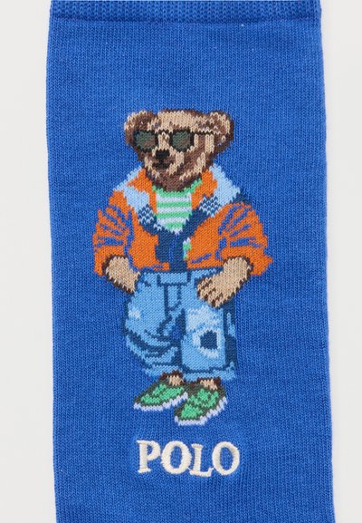 Πλεκτό μπλε ύφασμα με αρκουδάκι που φοράει γυαλιά ηλίου, πορτοκαλί μπουφάν, ριγέ πουκάμισο, μπλε τζιν, πράσινα παπούτσια και κείμενο «POLO» από κάτω.