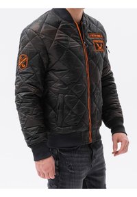 Vatteret bomberjakke i sort med orange detaljer. Har lynlås foran, ribbet ærmegab og patches på ærmerne. Materialet virker glat og skinnende.