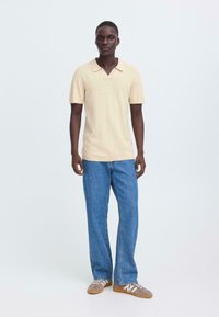 Polo beige à manches courtes avec un col en V, associé à un jean bleu ample et des baskets grises avec des bandes blanches.