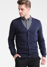 Mörkblå cardigan med V-halsdesign, med ribbad nederkant och ärmuddar, frontknäppning och lagd över en rutmönstrad skjorta.