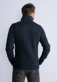 Maglione a maglia blu navy con colletto alto, caratterizzato da un motivo testurizzato e polsini e orlo a coste. Vista da dietro, indossato con pantaloni grigi.