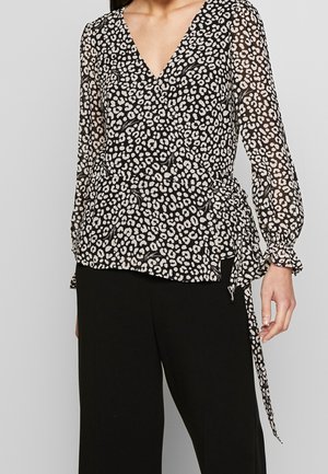 Kvinde iført sort og hvid leopardmønstret bluse med lange ærmer og bindedetalje, kombineret med ensfarvede sorte bukser med vide ben.