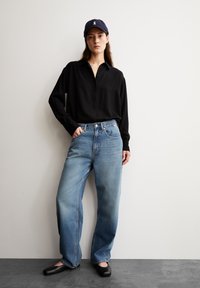 Camisa negra de manga larga, jeans de pierna ancha de lavado claro y bailarinas negras. El look incluye una gorra azul marino con un logo y un corte holgado en general.