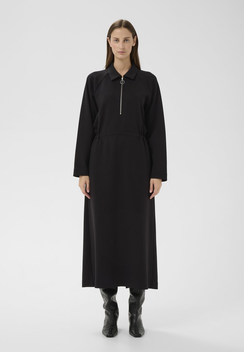 Mujer con un vestido largo negro de mangas largas, cuello, cremallera frontal y botas negras hasta la rodilla, de pie contra un fondo liso.