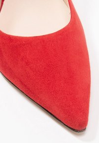 Scarpa rossa in suede con punta appuntita, caratterizzata da una texture liscia e una suola nera. Il design presenta una silhouette elegante e uno stile minimalista.