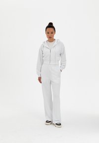 Felpa bianca corta con zip e coulisse, abbinata a pantaloni larghi a vita alta. L'outfit presenta un tessuto morbido e un design minimale.