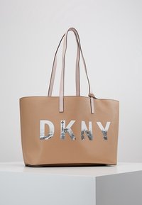 Sac fourre-tout beige avec longues poignées, arborant les lettres "DKNY" remplies d'une impression en niveaux de gris d'un paysage urbain, présenté sur un socle blanc contre un fond gris clair.