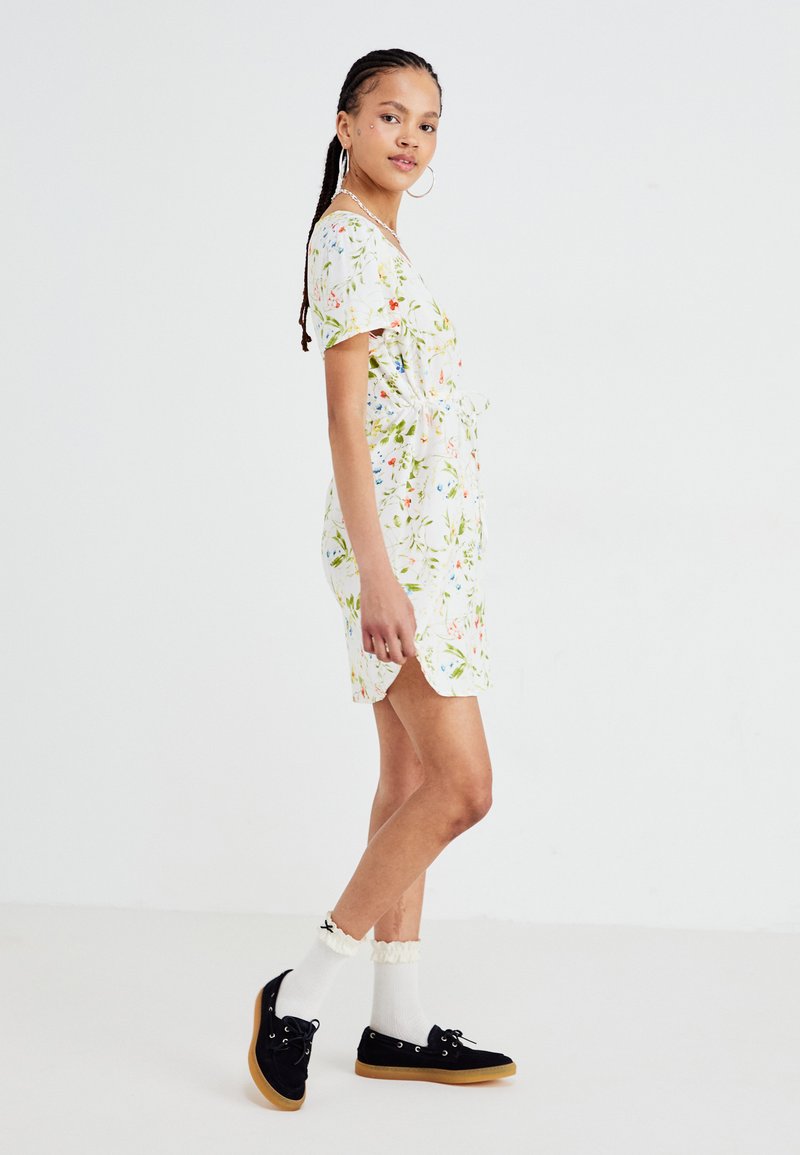 Blumenkleid kurz mit weißem Hintergrund, das grüne Blätter und bunte Blumen zeigt. Getragen mit schwarzen Schuhen und gerüschten weißen Socken.