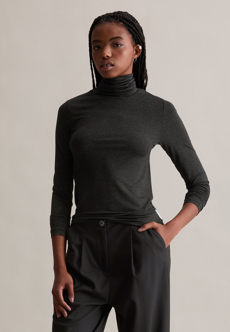 Schwarzer Rollkragenpullover mit langen Ärmeln, gefertigt aus einem schimmernden Stoff. Verfügt über ein figurbetontes Design und eine subtile Textur. Kombiniert mit schwarzen Hosen.