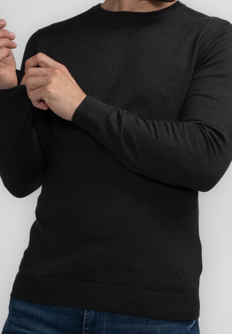 Homme ajustant la manche d'un pull noir à manches longues, portant un jean bleu, sur fond gris clair uni.