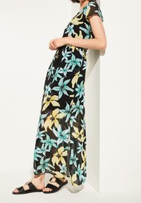Maxi-Kleid aus transparentem schwarzem Stoff mit großem Blumenmuster in Türkis und Gelb, mit kurzen Ärmeln und tailliertem Schnitt. Schwarze Sandalenflats.