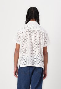 White patterned short-sleeve shirt made of eyelet fabric, featuring a collar and a straight hem. Back view reveals intricate cut-out design.