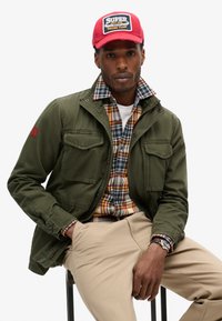 Zielona kurtka z płótna z czterema przednimi kieszeniami, noszona na koszuli w kratę. Czerwona czapka z logo, w zestawie z beżowymi spodniami i zegarkiem na rękę.