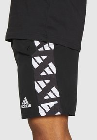 Pantalones cortos deportivos negros con un logo blanco de Adidas y una banda geométrica blanca en el lateral, llevados por una persona de piel oscura.