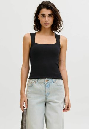 Göndör hajú nő fekete bordázott ujjatlan felsőt és világoskék magasderekú farmert visel, sima háttér előtt áll.