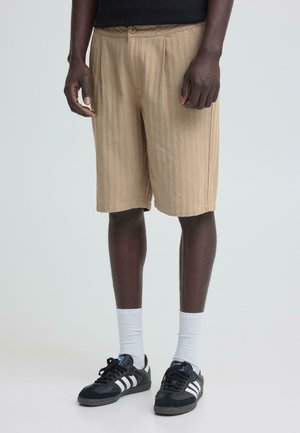 Uomo con pantaloncini beige fino al ginocchio, sneaker nere con strisce bianche, calzini bianchi a costine, in piedi davanti a uno sfondo semplice.