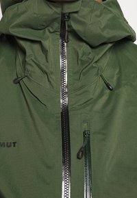 Zielona kurtka na zewnątrz z wysokim kołnierzem, wyposażona w centralny zamek błyskawiczny, dwie boczne kieszenie oraz regulowane sznurki umożliwiające dopasowanie na ciasno.