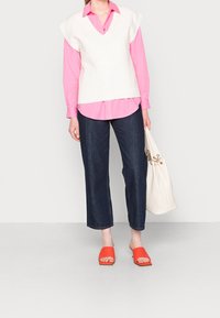 Vit stickad väst med v-ringning över en rosa knapp-up skjorta, i kombination med lösa mörk denimbyxor och klara orangea fyrkantiga mules.