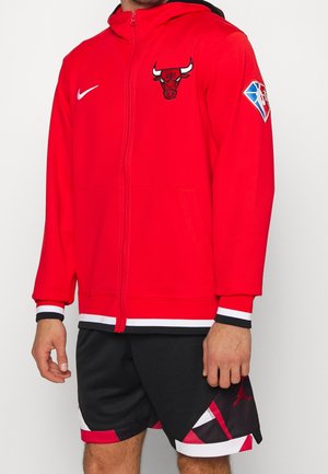 Uomo che indossa una giacca con zip rossa dei Chicago Bulls con loghi e pantaloncini da basket neri con dettagli rossi e bianchi.