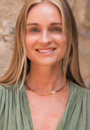 Gold chain necklace featuring rectangular links, worn with a green textured top. Background is blurred, suggesting a natural outdoor setting.