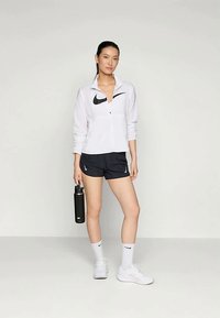 Ženska v beli jakni na zips in črnih kratkih hlačah, ki drži črn steklenico za vodo, stoji ob svetli enobarvni ozadju.