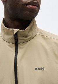 Άνδρας που φοράει μπεζ μπουφάν με φερμουάρ και ψηλό γιακά, με μαύρο λογότυπο "BOSS" στο στήθος.