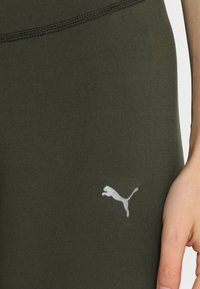 Zelené sportovní legíny vyrobené z elastického materiálu, s bílým logem Puma na dolní levé straně a hladkým, matným povrchem.