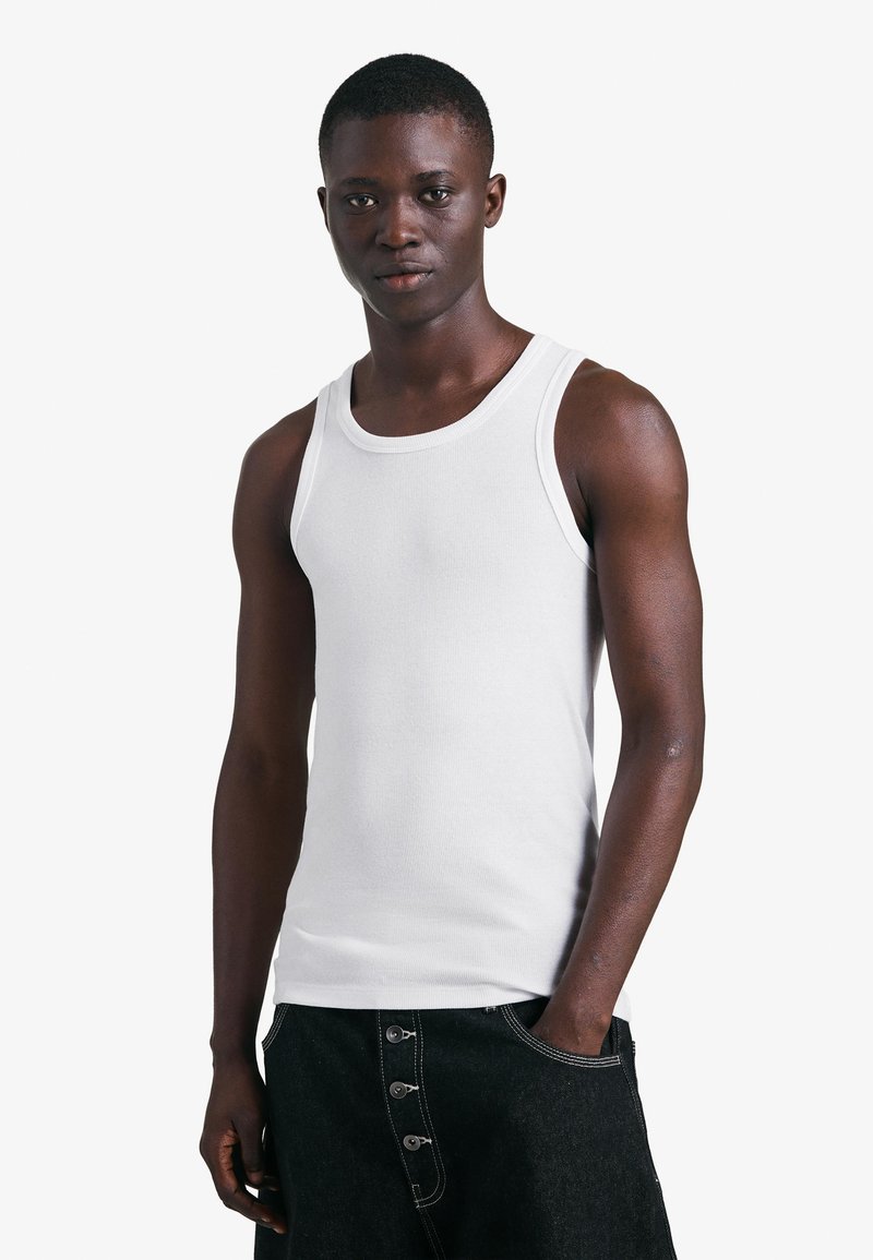 Mlad moški, oblečen v belo majico brez rokavov in črne hlače z gumbi, stoji z eno roko v žepu na belem ozadju.