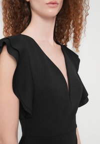 Γυναίκα με κατσαρά κόκκινα μαλλιά που φοράει μαύρο φόρεμα με βαθύ V ντεκολτέ και κοντά μανίκια με βολάν, εμφανιζόμενη από τους ώμους έως τη μέση.