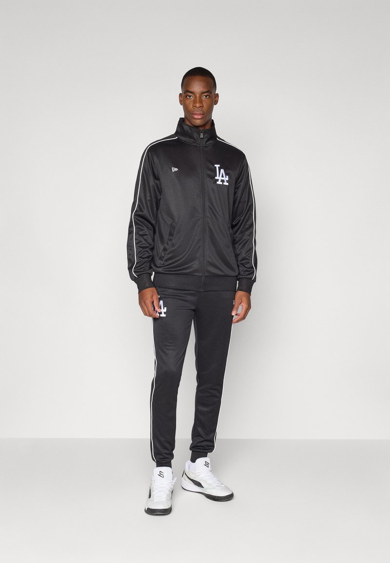 Black zip-up tracksuit made of shiny fabric, featuring white stripes on sleeves and sides, with LA logo on chest and pants. White sneakers.