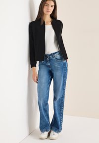 Giacca nera con cerniera e texture a coste, indossata sopra una maglietta bianca; jeans larghi a gamba larga azzurri con finitura slavata; sneaker bianche.