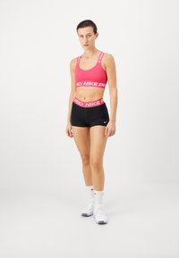 Różowy stanik sportowy z białym logotypem "Nike Pro" i dopasowanym pasem, w połączeniu z czarnymi szortami. Białe, niskie buty sportowe z fioletowymi akcentami.