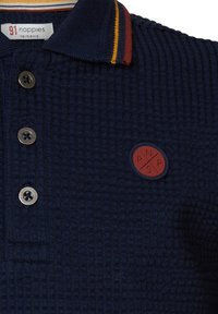 Dunkelblaues Polohemd aus strukturiertem Gewebe, mit drei Knopfverschluss, einem gestreiften Kragen in Gelb und Rot sowie einem runden Gummiemblem.