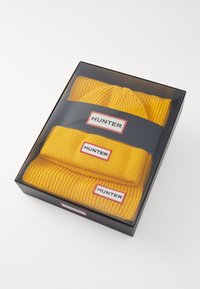Žuta pletena kapa i odgovarajući šal, predstavljeni u prozirnoj kutiji. Sadrže oznake "HUNTER" na oba predmeta u kontrastnoj crvenoj i bijeloj boji.