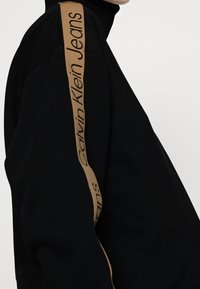Sudadera negra con cuello alto, que presenta una franja de logotipo marrón a lo largo del hombro, fabricada en un tejido suave y texturizado.