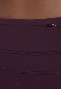 Zbliżenie na ciemnofioletowe legginsy sportowe z gładkim pasem i małą czarną kieszonką na zamek z boku.