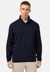 Jūras flotes kreklis ar apkakli, teksturēts audums, poga plaukts un garas piedurknes. Novietots uz neitrāla fona.