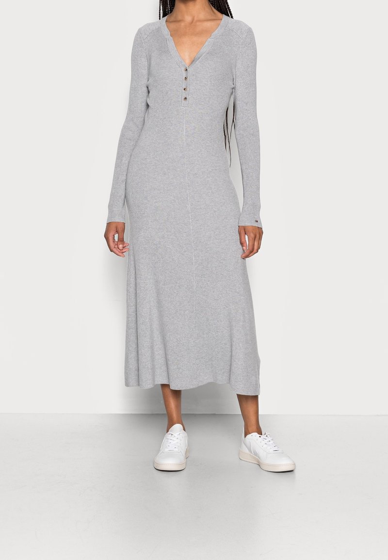 Femme portant une longue robe en maille côtelée grise avec des boutons sur le col en V, associée à des baskets blanches, debout sur un fond uni.