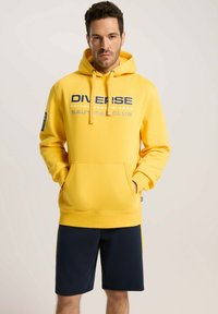 Żółta bluza z kapturem, z kieszenią na froncie, ozdobiona wyraźnym tekstem i logo, w połączeniu z ciemnonawigacyjnymi szortami. Miękki materiał i swobodny krój.