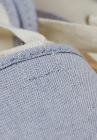 Pantofle acoperite cu țesătură de culoare albastru deschis, cu o suprafață texturată, evidențiind cusătura în contrast și o curea din pânză. Vedere de aproape care pune accent pe detaliile cusăturii.