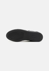 Suola nera di scarpe con motivi testurizzati per la presa, caratterizzata da scanalature parallele e un emblema circolare. Realizzata in gomma con un profilo piatto.
