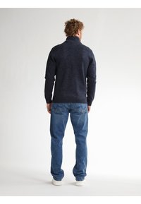 Marineblauer Strickpullover mit hohem Kragen, Rippdetails an den Schultern, kombiniert mit hellblauen Jeans und weißen Sneakers.