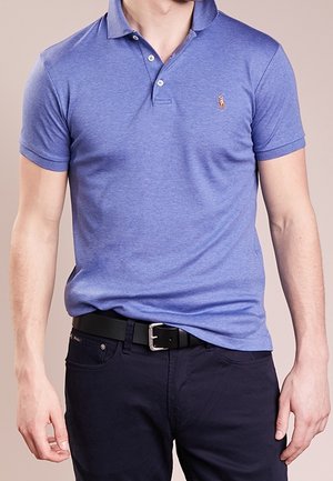 Man wearing a fitted blue polo shirt with a small embroidered logo, dark pants, and a black belt with a silver buckle.